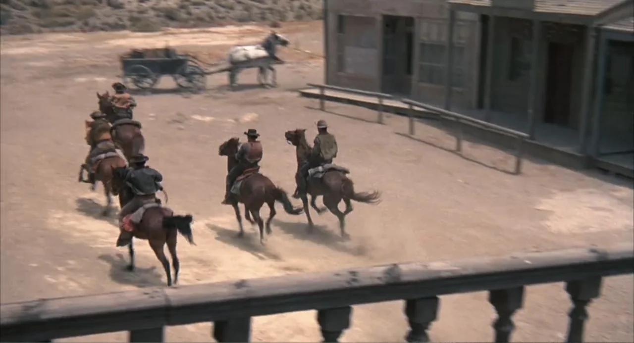 The Spikes Gang (1974) movie still: wide — A dusty dirt street in a wooden western town sits under a bright sky. Five men on horseback travel past a…