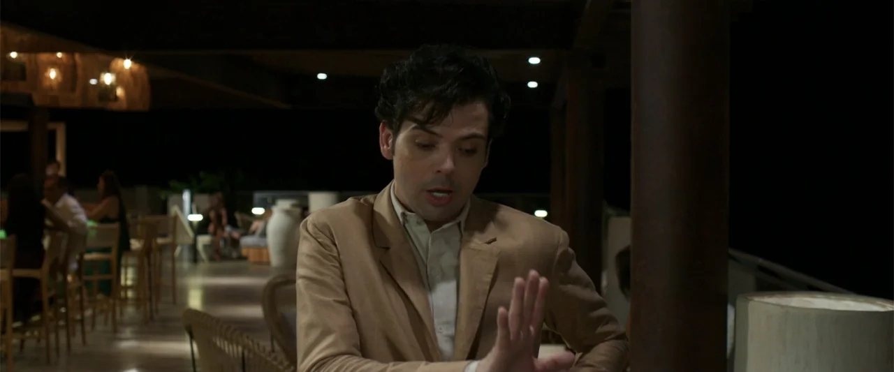 Férias Trocadas (2024) movie still: medium — An outdoor bar terrace at night features dark wooden architectural pillars and rattan furniture. A man…