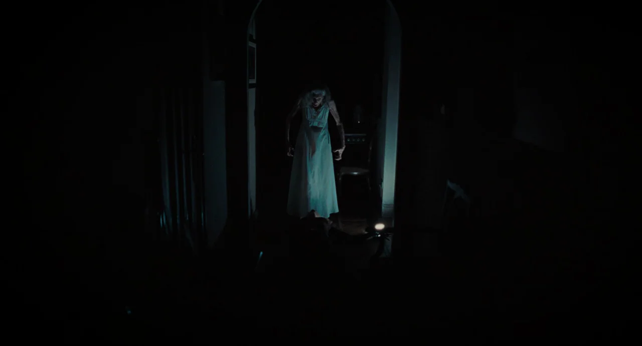 La Abuela (2021) movie still: wide — A dark hallway leads to an arched doorway containing a pale, standing woman. The woman in the white nightgown…