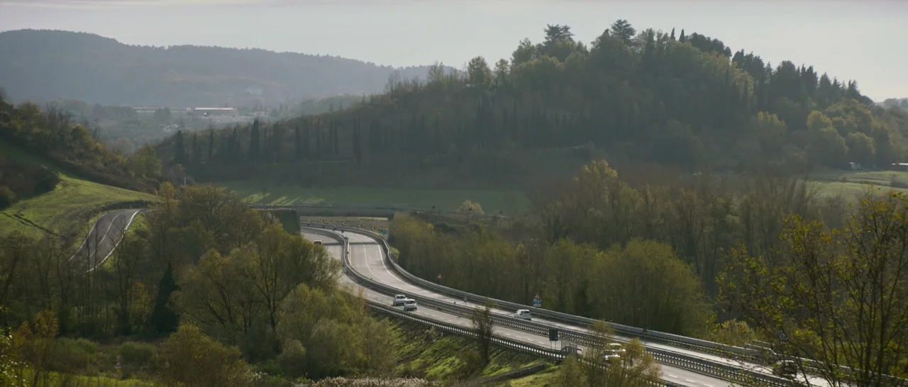 The Face of an Angel (2014) movie still: establishing — An asphalt highway winds through a rolling green valley filled with deciduous trees. Hills…