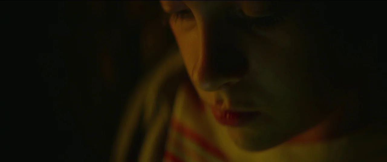 Scrapper (2023) movie still: closeup — A close-up of a child in a dark space, illuminated by a dim light. The subject looks downward with eyes…