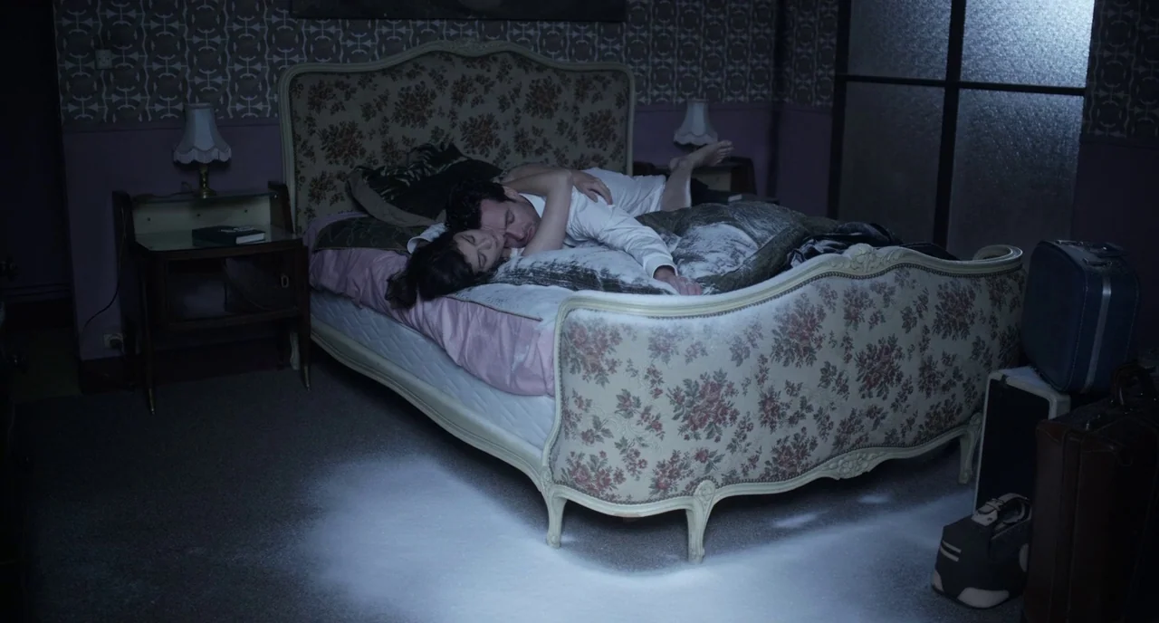 Mood Indigo (2013) movie still: wide — An dimly lit, old-fashioned hotel room features patterned wallpaper and a vintage, floral-upholstered bed. A…
