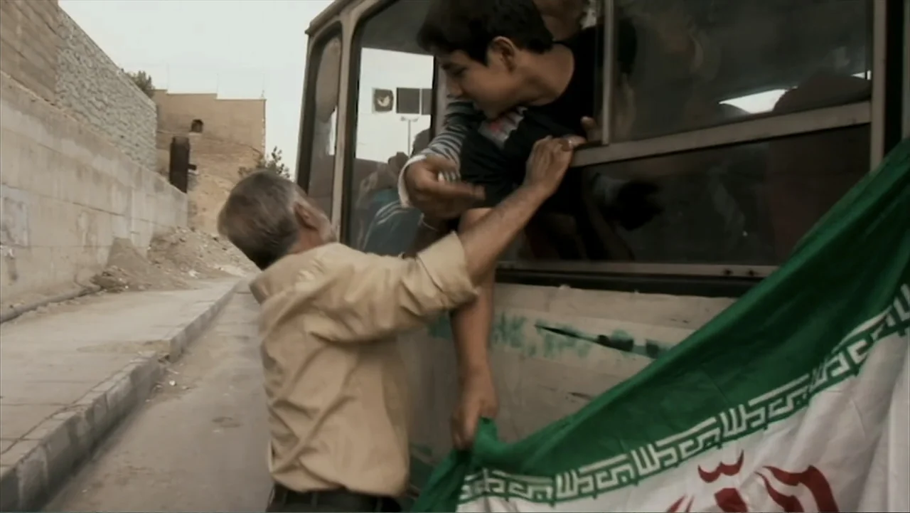 Offside (2015) movie still: medium — A street scene featuring a large vehicle with a flag draped over the side. An older man leans toward a young…