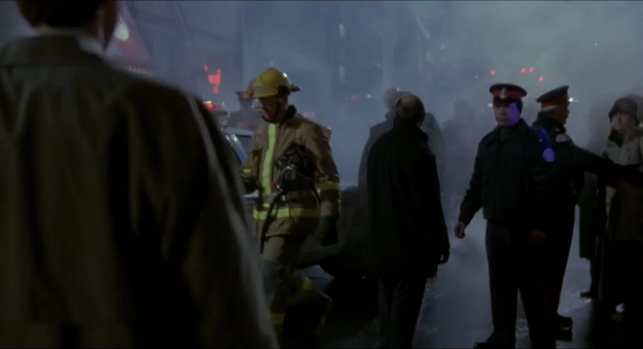 Foolproof (2003) movie still: medium — A dark city intersection is filled with thick smoke and emergency vehicle lighting. A firefighter walks past…