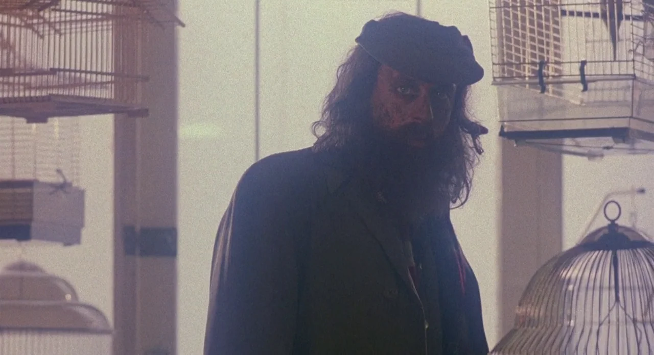 Hellraiser (1987) movie still: medium — A man with a thick beard and cap stands amidst hanging metal birdcages. The background is a bright,…