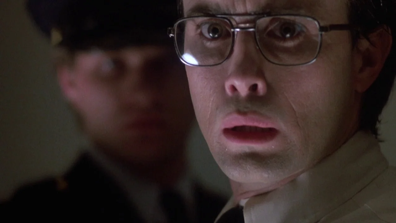 Re-Animator (1985) movie still: closeup — A dimly lit interior space with two men close together. One man in glasses looks directly ahead with wide…