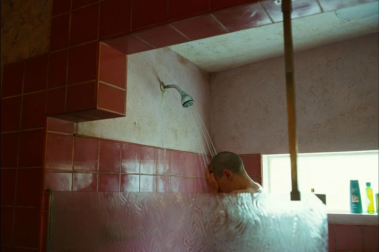 Bugonia (2025) movie still: medium — A residential shower stall features worn mauve tiles and a textured plastic curtain. A person with a shaved…