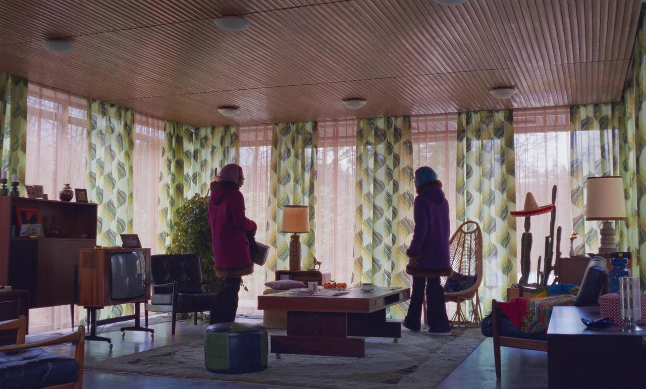 The Silent Twins (2022) movie still: wide — A 1970s style living room features patterned olive and cream floor-to-ceiling curtains. Two women…