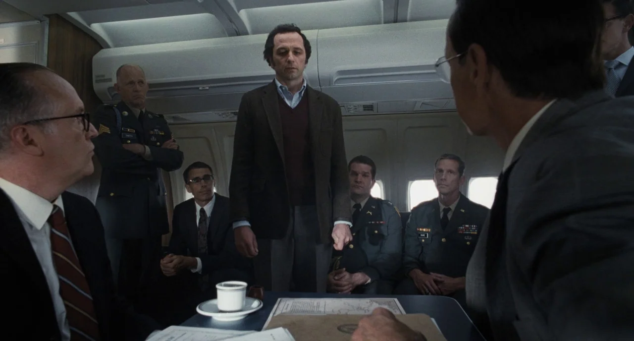 The Post (2017) movie still: medium — A group of men in suits and military uniforms sit inside an aircraft cabin. A man stands in the center aisle…