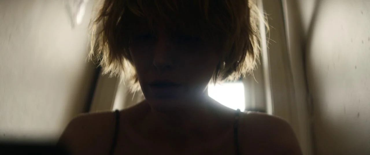 The Rhythm Section (2020) movie still: closeup — A dimly lit interior space with a bright light source located directly behind the subject. A woman…