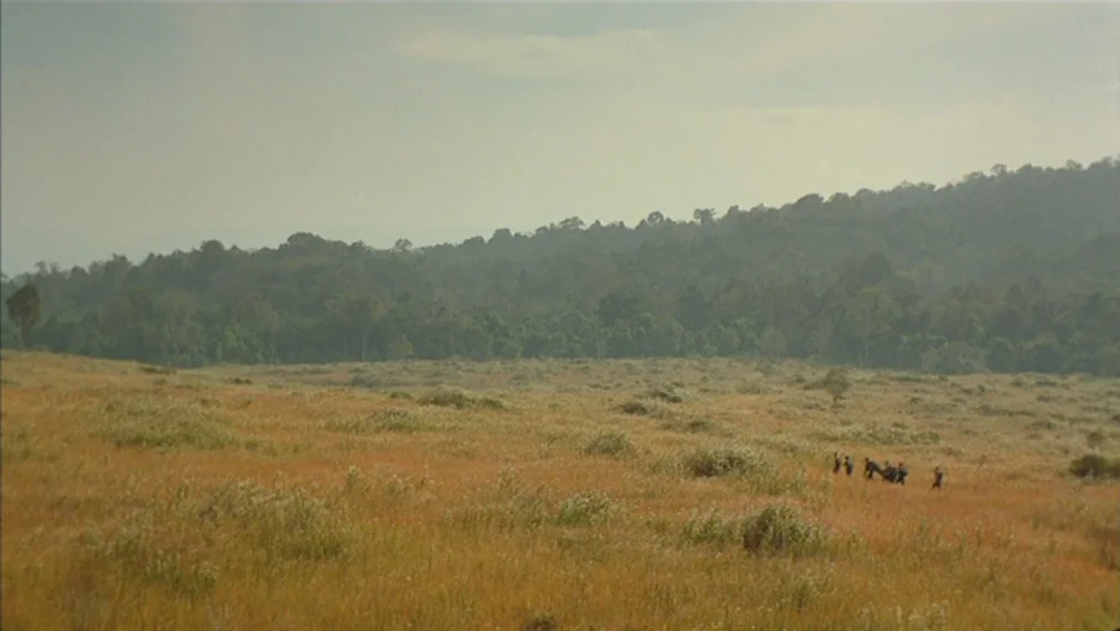 Tropical Malady (2004) movie still: wide — A vast, dry grassy plain stretches toward a dense tree line under a pale sky. A small group of indistinct…