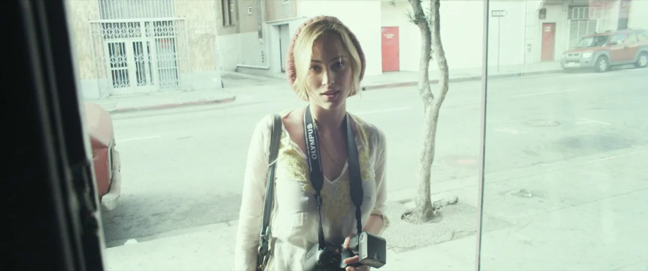 Maniac (2012) movie still: medium — A city sidewalk with a bare tree and buildings in the background. A young blonde woman stands facing the camera…