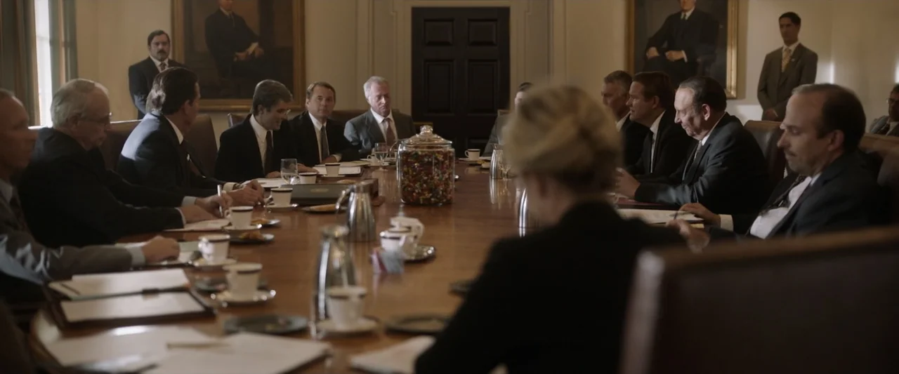 Reagan (2024) movie still: wide — A formal meeting occurs in a wood-paneled room with large framed portraits on walls. Multiple men in business…