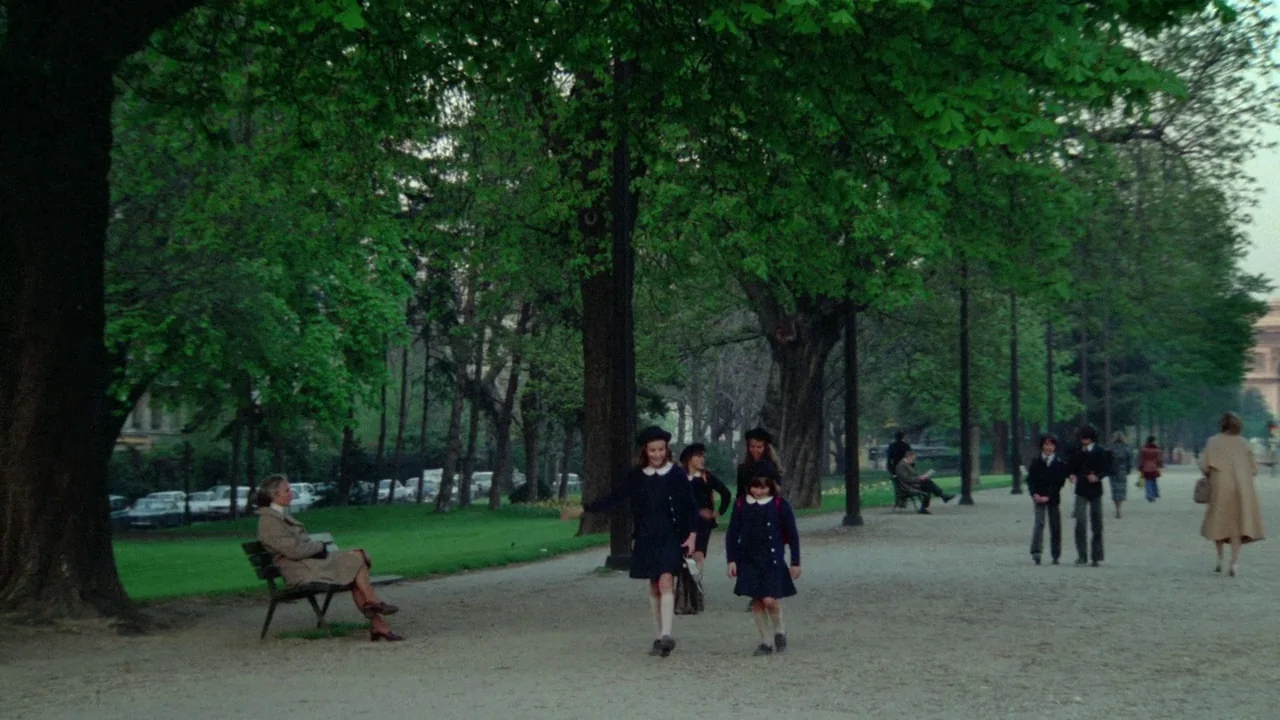 Sorcerer (1977) movie still: wide — A public park features a wide gravel path shaded by large mature deciduous trees. Schoolgirls in navy uniforms…