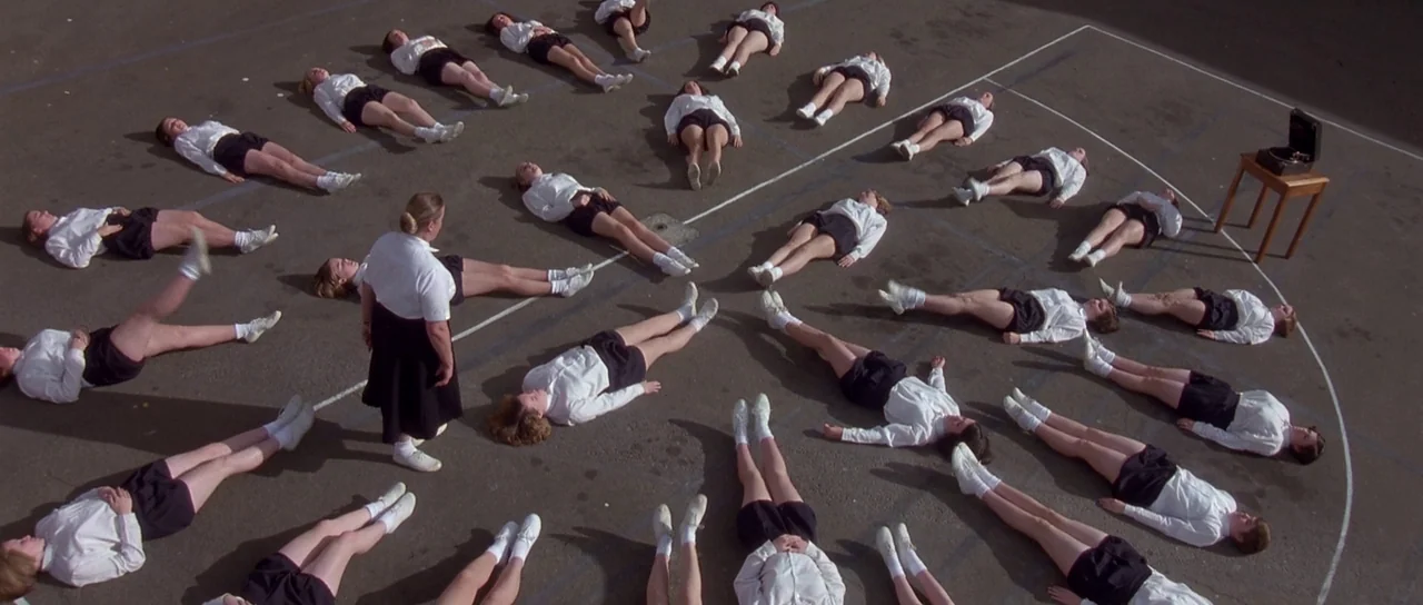 Heavenly Creatures (1994) movie still: wide — An asphalt surface is marked with white lines in a large empty lot. A group of people in matching…