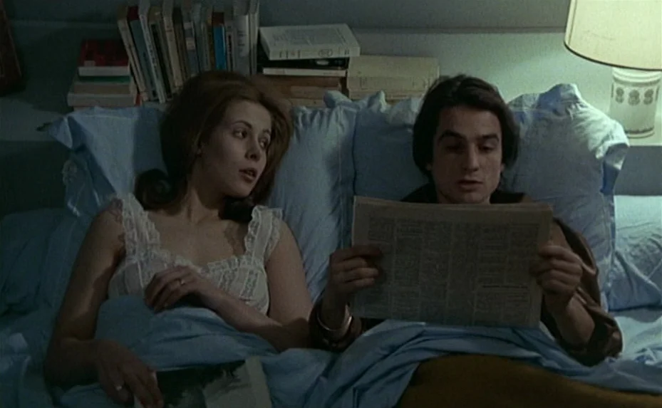 Love on the Run (1979) movie still: medium — A man and woman lie together in a bed covered with light blue sheets. A stack of books sits on the…