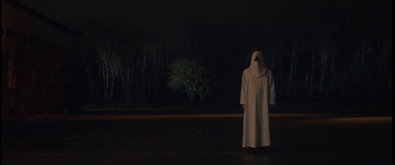 The Void (2016) movie still: fullbody — A dark empty asphalt lot is bordered by a line of bare trees at night. A lone figure dressed in a white robe…