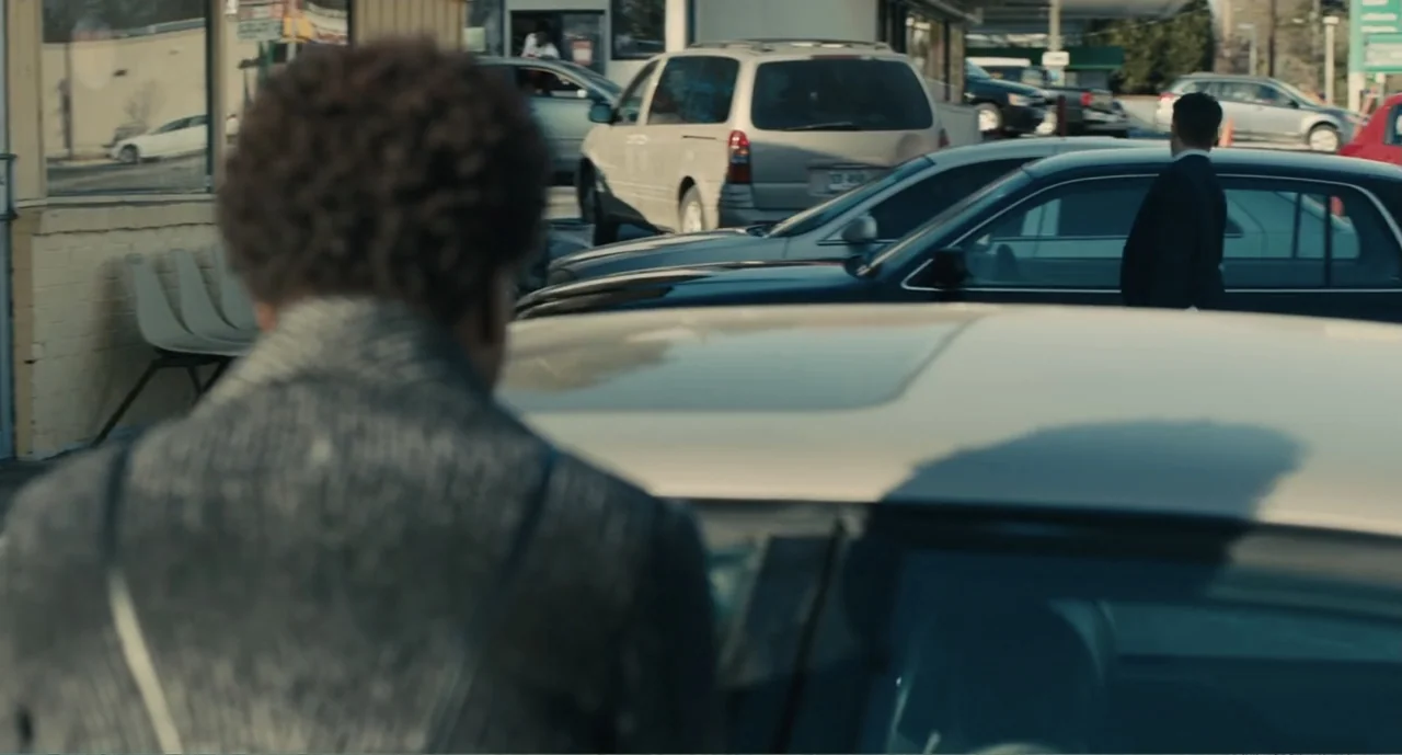 Lila & Eve (2015) movie still: medium — A sunny parking lot filled with various modern cars during the day. A man stands in the foreground facing a…