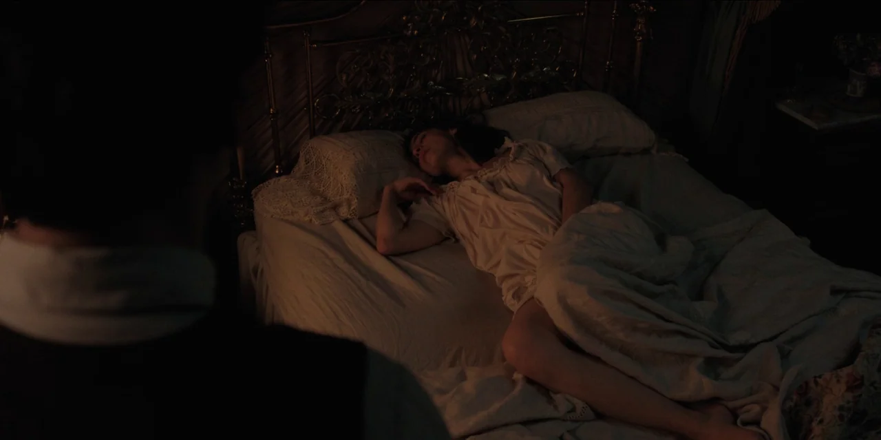 Pedro Páramo (2024) movie still: medium — A dark, wood-paneled room features a vintage brass bed frame with lace-trimmed linens. A woman sleeps on…