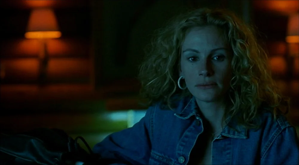Erin Brockovich (2000) movie still: medium — A dimly lit indoor space with blurred lamps in the background. A woman sits in the foreground, facing…