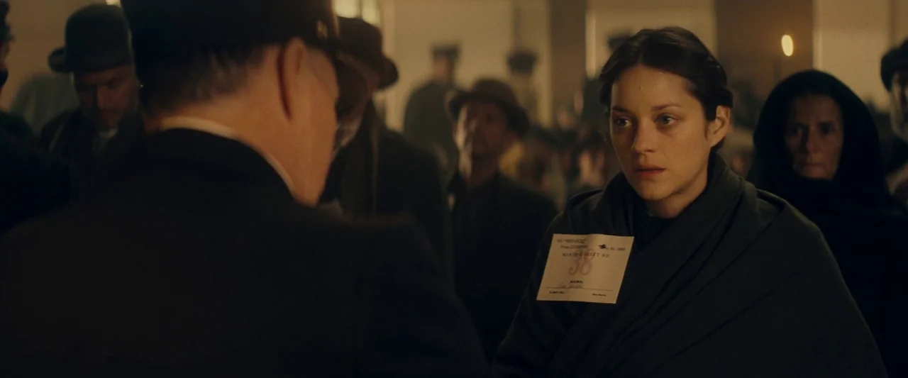 The Immigrant (2013) movie still: medium — A crowded hall with many people waiting in line wearing vintage hats. A woman looks forward while wearing…
