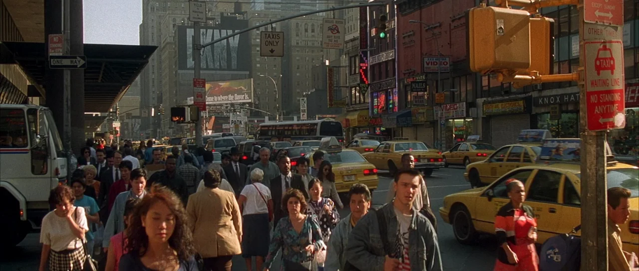 Die Hard with a Vengeance (1995) movie still: wide — A dense metropolitan street intersection crowded with pedestrians and vintage 1990s yellow…
