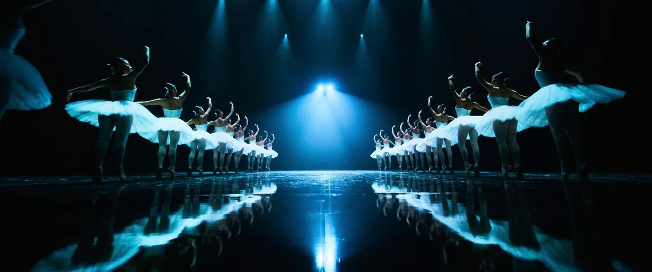 Ballerina (2023) movie still: wide — A dark theater stage features two long rows of ballerinas facing toward the back. The glossy black floor…