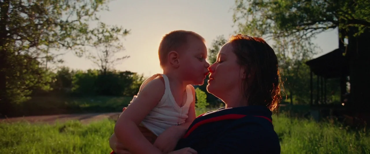 Lost River (2014) movie still: medium — A sun-drenched grassy meadow features large trees under a late afternoon sky. A woman holds a young child in…