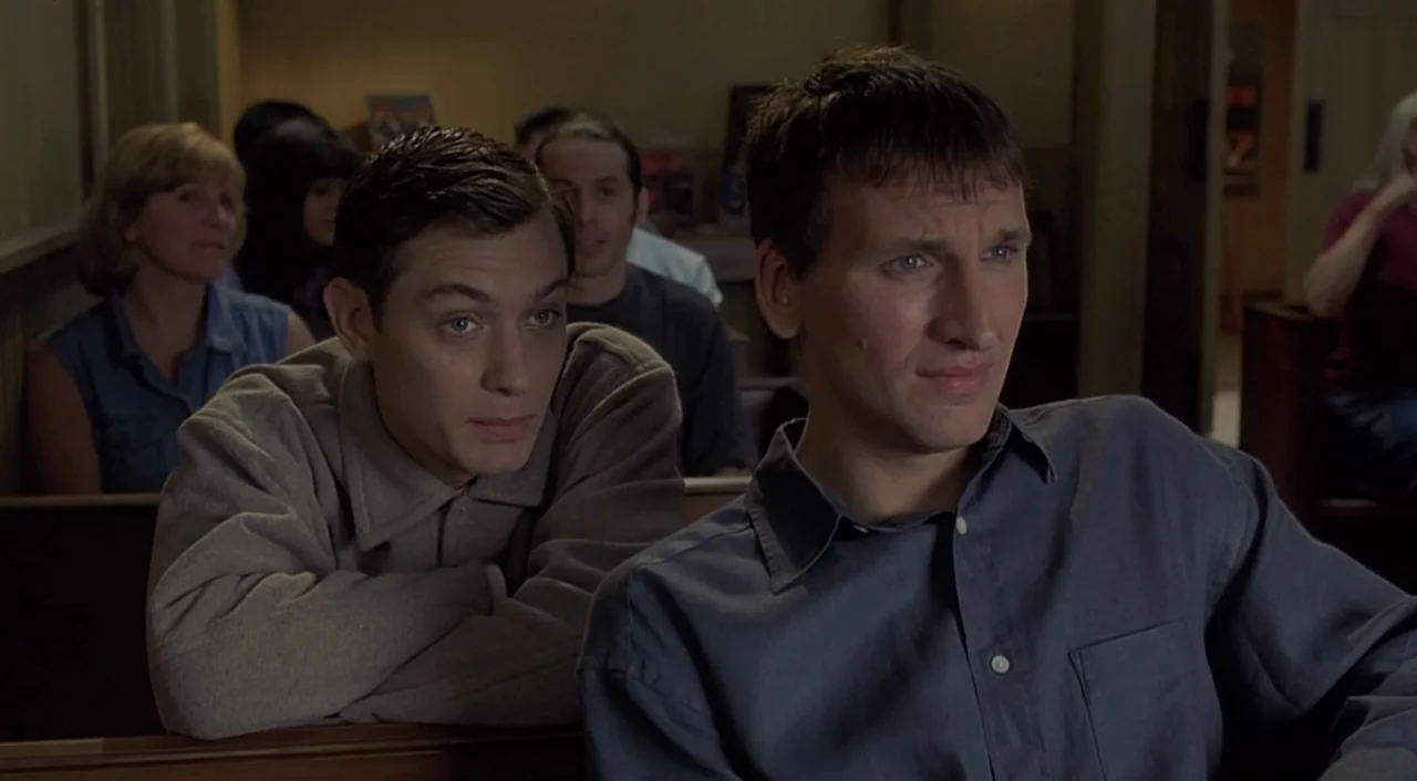 eXistenZ (1999) movie still: medium — An interior space with wooden benches arranged in rows for an audience. Two men sit in the front row watching…