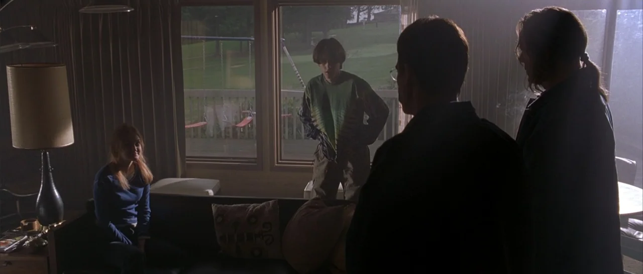 Bandits (2001) movie still: medium — A dimly lit residential living room with a large window looking onto a lawn. Three young adults stand and sit…