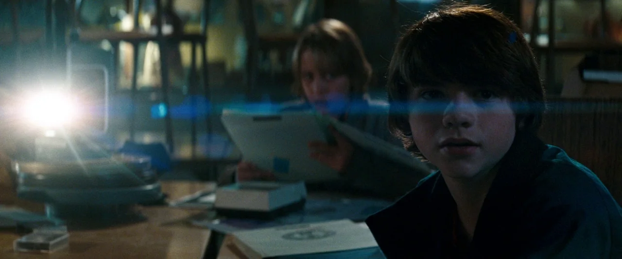 Super 8 (2011) movie still: medium — A dimly lit basement features a work table covered in documents and a projector. Two teenagers are seated at…