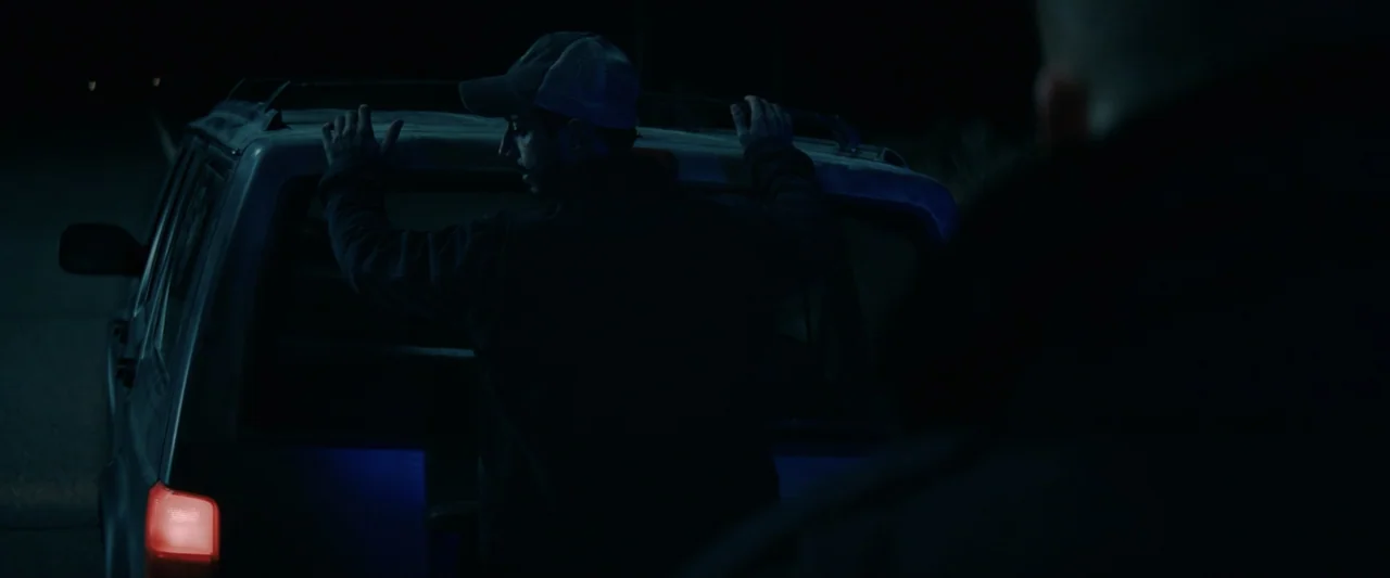 Encounter (2018) movie still: medium — A dark outdoor space at night shows a man leaning against a vehicle trunk. The man remains in shadows while…