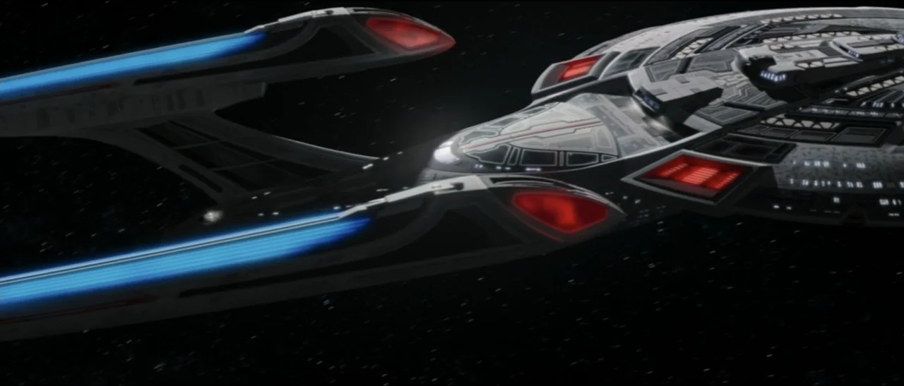 Star Trek: Nemesis (2002) movie still: wide — A complex starship with elongated blue nacelles and a grey saucer section traverses deep space. The…