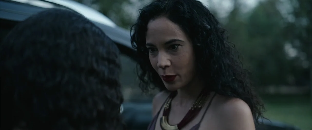 Evil Eye (2020) movie still: medium — A dark outdoor space features a woman in the background facing another person. The woman has long dark hair…