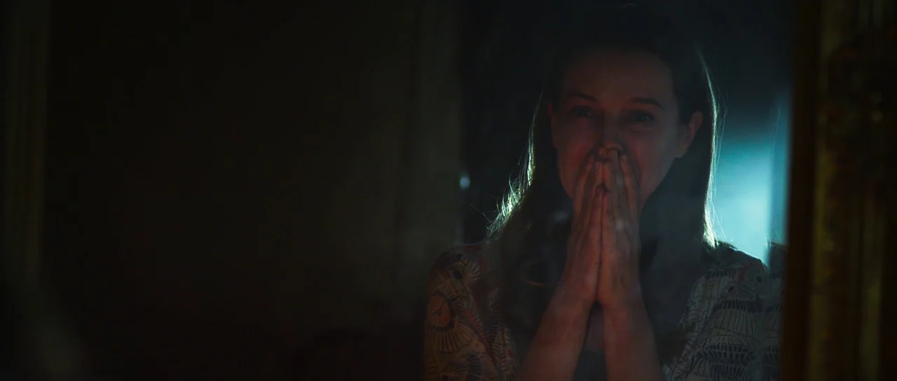 A Dark Song (2016) movie still: closeup — A dark interior space with a person visible behind a reflective glass surface. The woman stands with both…