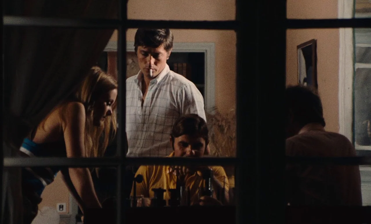 The Swimming Pool (1969) movie still: medium — A group gathers inside a room at night viewed through a grid window. Three people look toward a man…