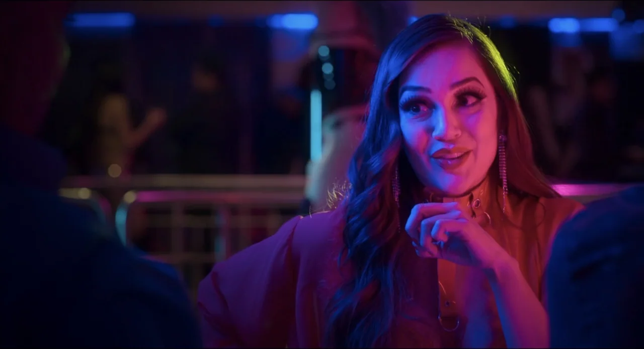 The Beauty and the Moody (2022) movie still: medium — A dimly lit nightclub interior features neon lighting and a metal railing background. A woman…