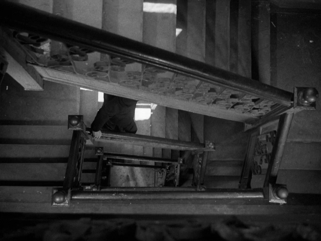 The Naked City (1948) movie still: wide — An ornate interior staircase features intricate metal railings and dark wooden steps. A person in a suit…