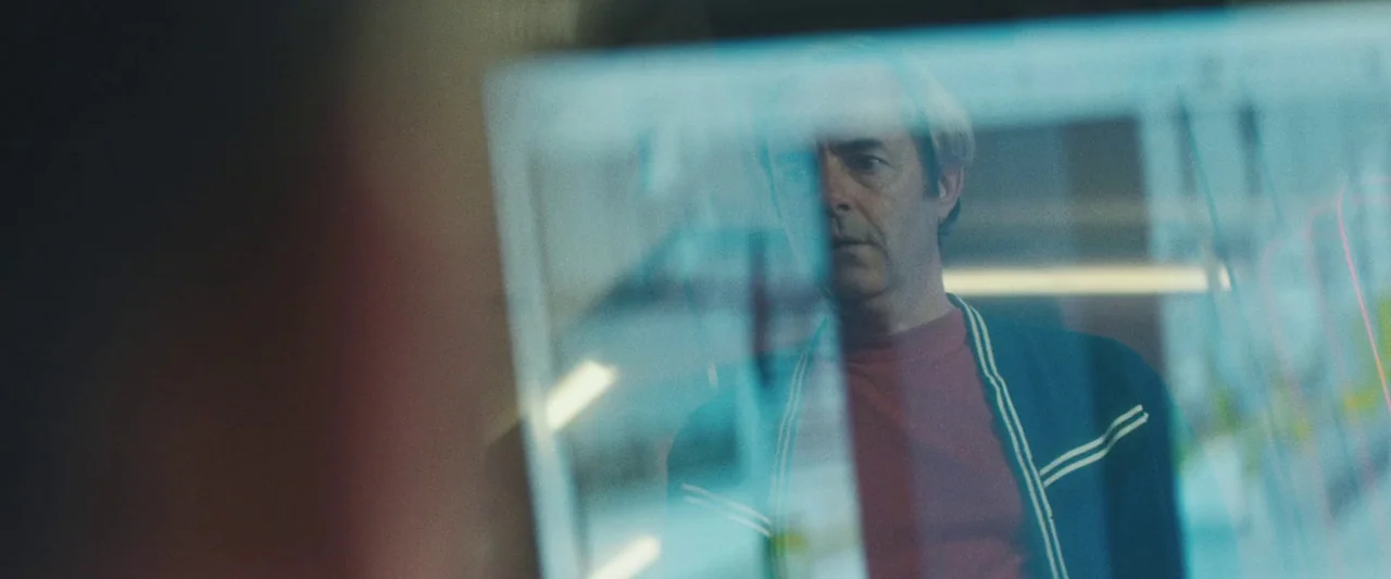 Vincent Must Die (2023) movie still: medium — A man stands behind a glass partition inside a brightly lit office space. The reflection of the…