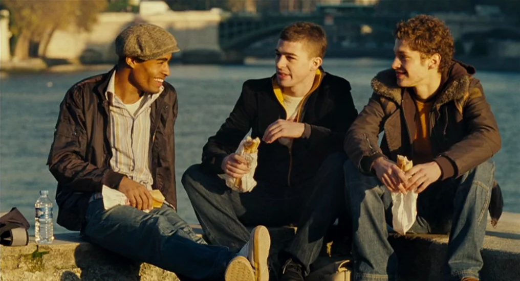 Paris, Je T’Aime (2006) movie still: fullbody — Three young men sit on a stone wall beside a wide river at dusk. They hold paper-wrapped sandwiches…