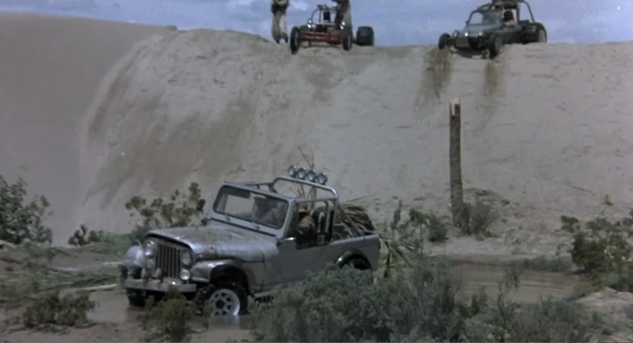 Firewalker (1986) movie still: wide — A silver jeep sits in a muddy pool at the base of a large sand dune. Two sand buggies descend the crest of the…