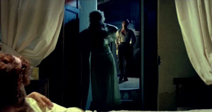 Blood For Dracula (1974) movie still: medium — A woman stands in a shadowed doorway looking toward a man in the distance. The man stands in a dimly…