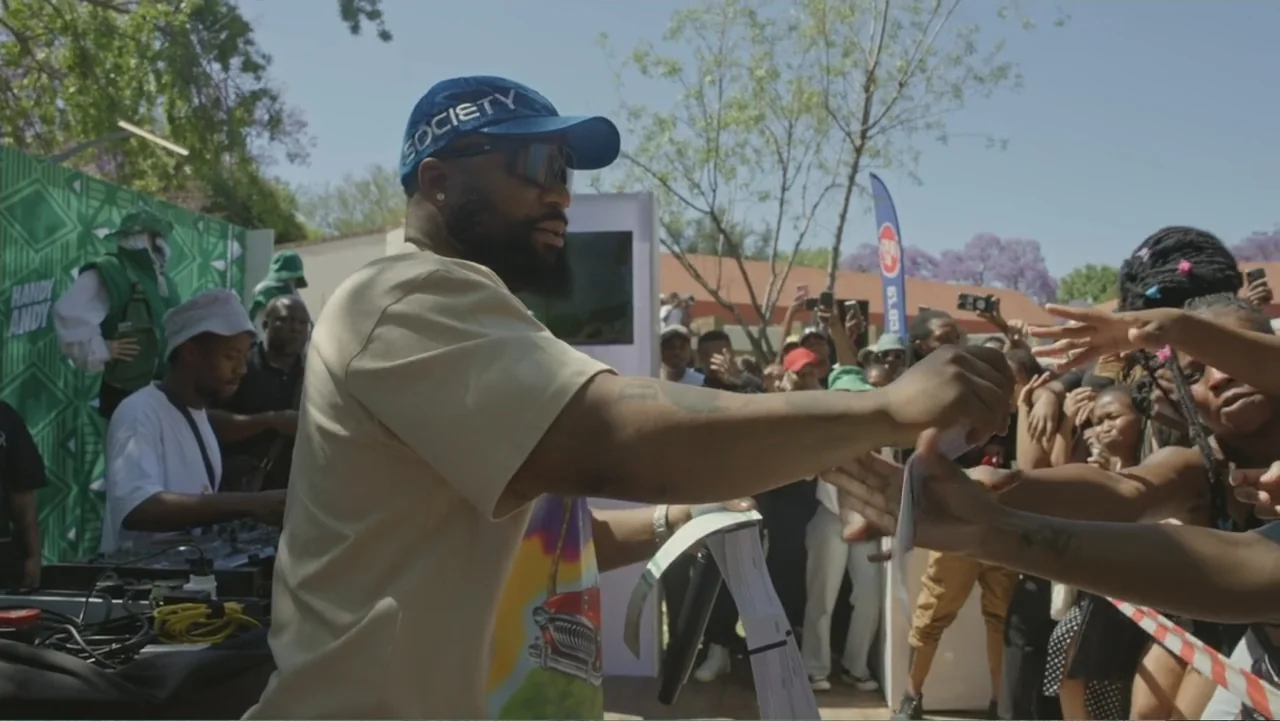 African Throne World Tour (2024) movie still: medium — An outdoor event features a performer at a booth in front of a crowd. The man hands out…
