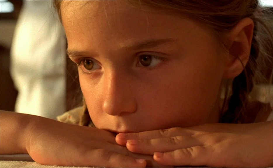 Chocolat (1988) movie still: closeup — A close-up view of a young child in a sunlit room. She rests her chin on her hands while looking towards the…