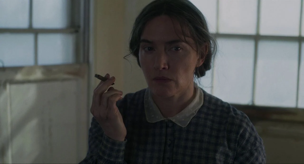 Ammonite (2020) movie still: medium — A woman stands in a plain room with large grid-paned windows in the background. She holds a cigar in her hand…