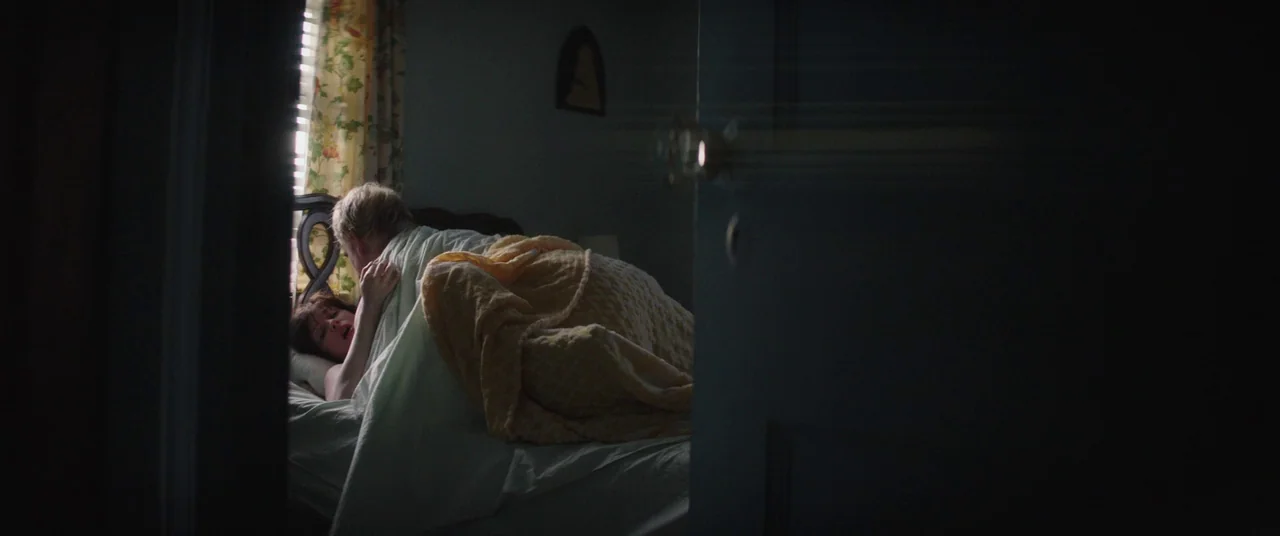 God’s Pocket (2014) movie still: wide — A dark, partially obscured view into a room from behind a door frame. A man and woman rest together in bed…