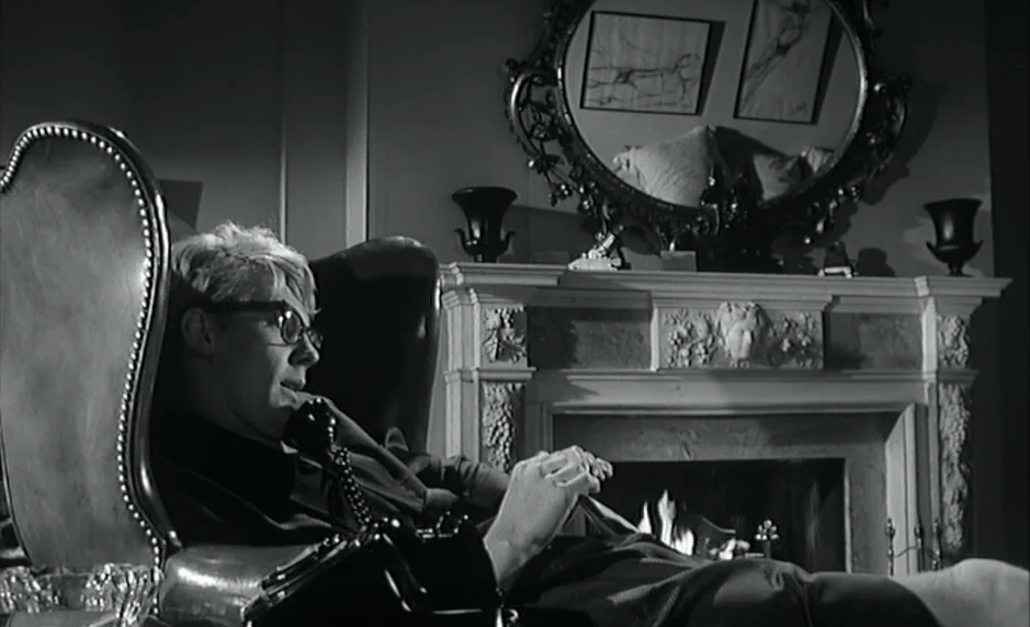 The Servant (1963) movie still: medium — A dark living room features a carved marble fireplace with a large decorative mirror. A young man sits in…