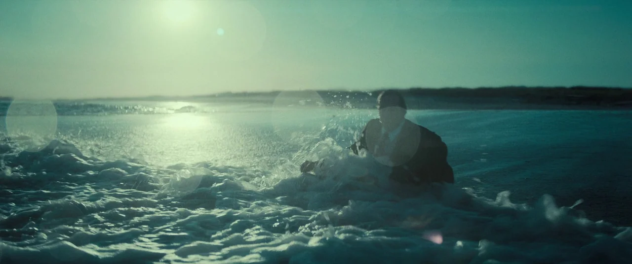 The Many Saints of Newark (2021) movie still: wide — A vast, flat sandy beach stretches toward the horizon under a bright sun. A man in a suit sits…