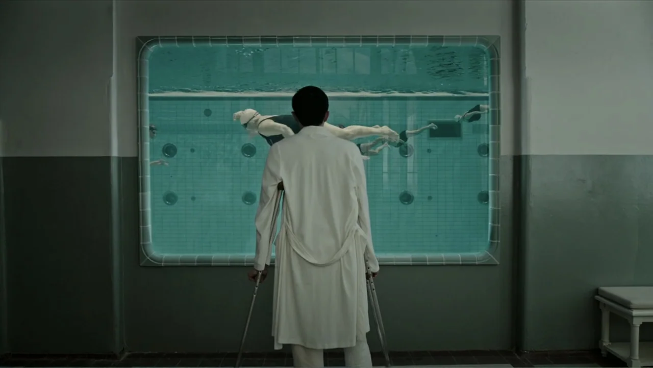 A Cure for Wellness (2017) movie still: fullbody — A clinical tiled therapy facility features a large reinforced glass window into a pool. A man in…