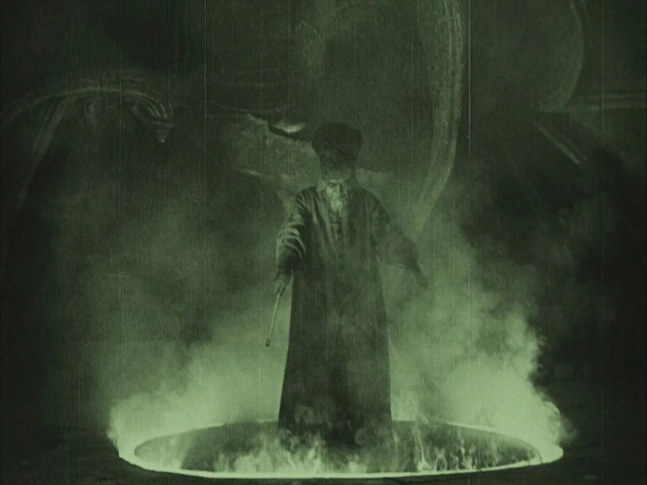 The Golem (1920) movie still: fullbody — A stone chamber features carved walls behind an elderly man standing inside a fire circle. The figure…